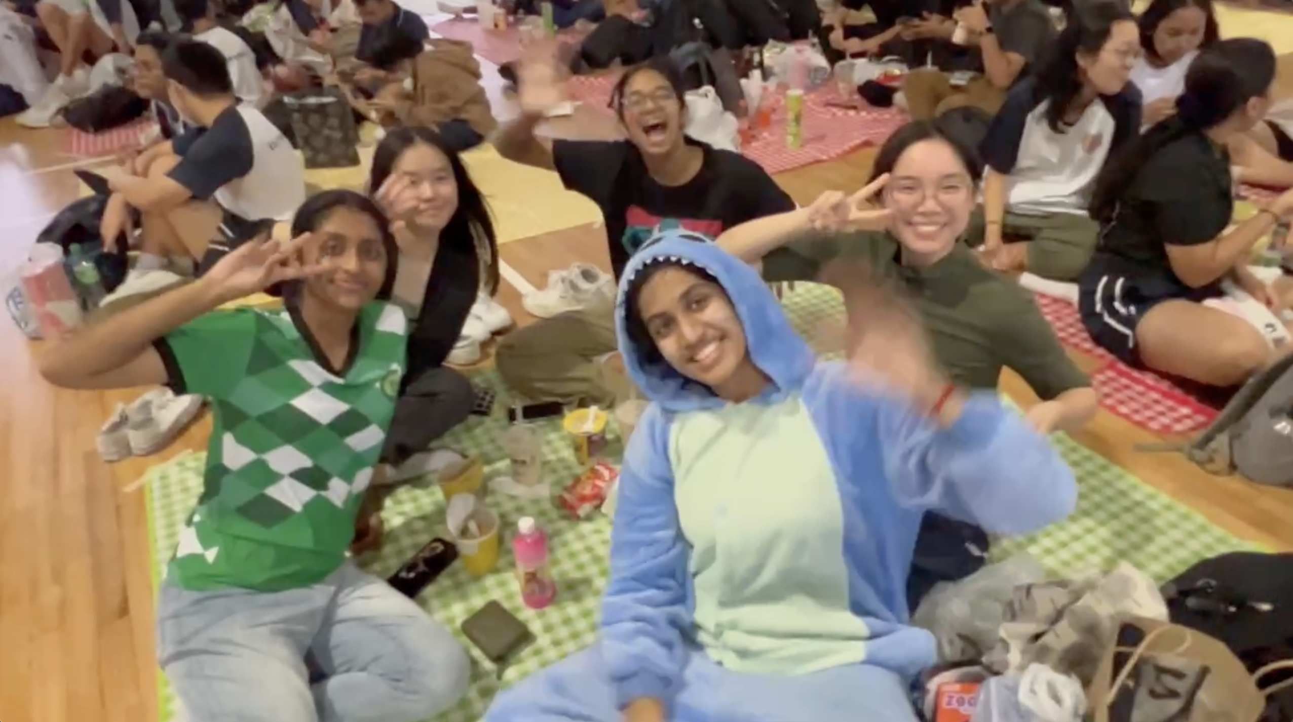 Five friends sit on picnic mats indoors, smiling and waving; one wears a blue onesie, others flash peace signs.
